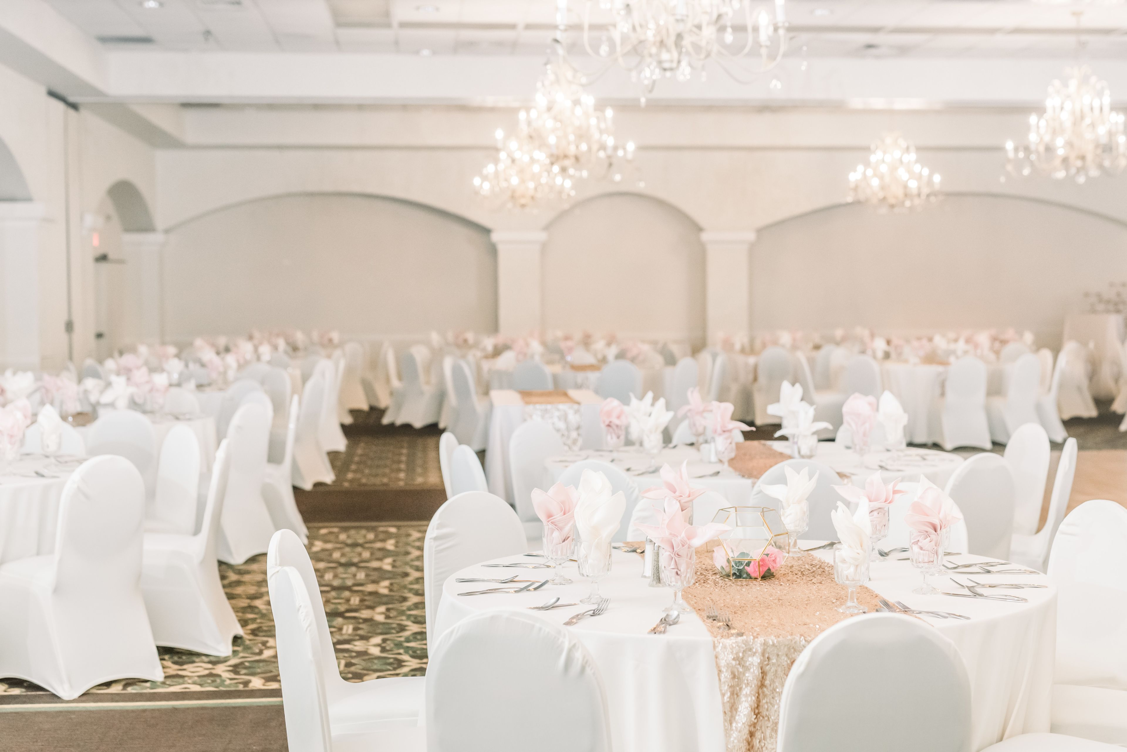 Une salle de banquet avec des tables rondes dressées pour un événement formel, agrémentée de nappes et de chaises blanches, et de lustres suspendus au plafond.