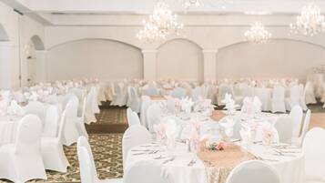 Une salle de banquet avec des tables rondes dressées pour un événement formel, agrémentée de nappes et de chaises blanches, et de lustres suspendus au plafond.