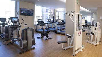 Una palestra con diverse attrezzature per cardio e allenamento della forza, ampie finestre e un televisore a schermo piatto sulla parete.