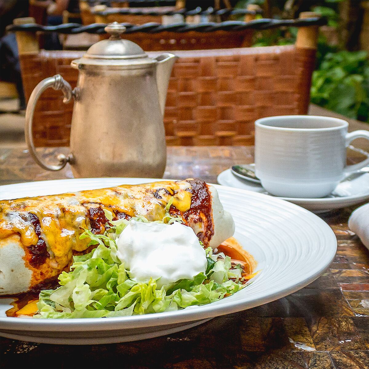 A plate of enchiladas with sour cream and a side salad, a white cup, and a metal pitcher on a table.