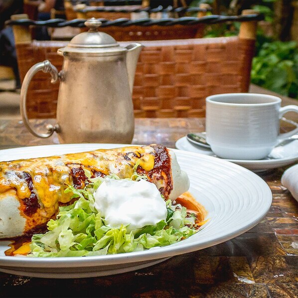 Un plato de enchiladas con crema agria y una ensalada, una taza blanca y una jarra de metal sobre una mesa.
