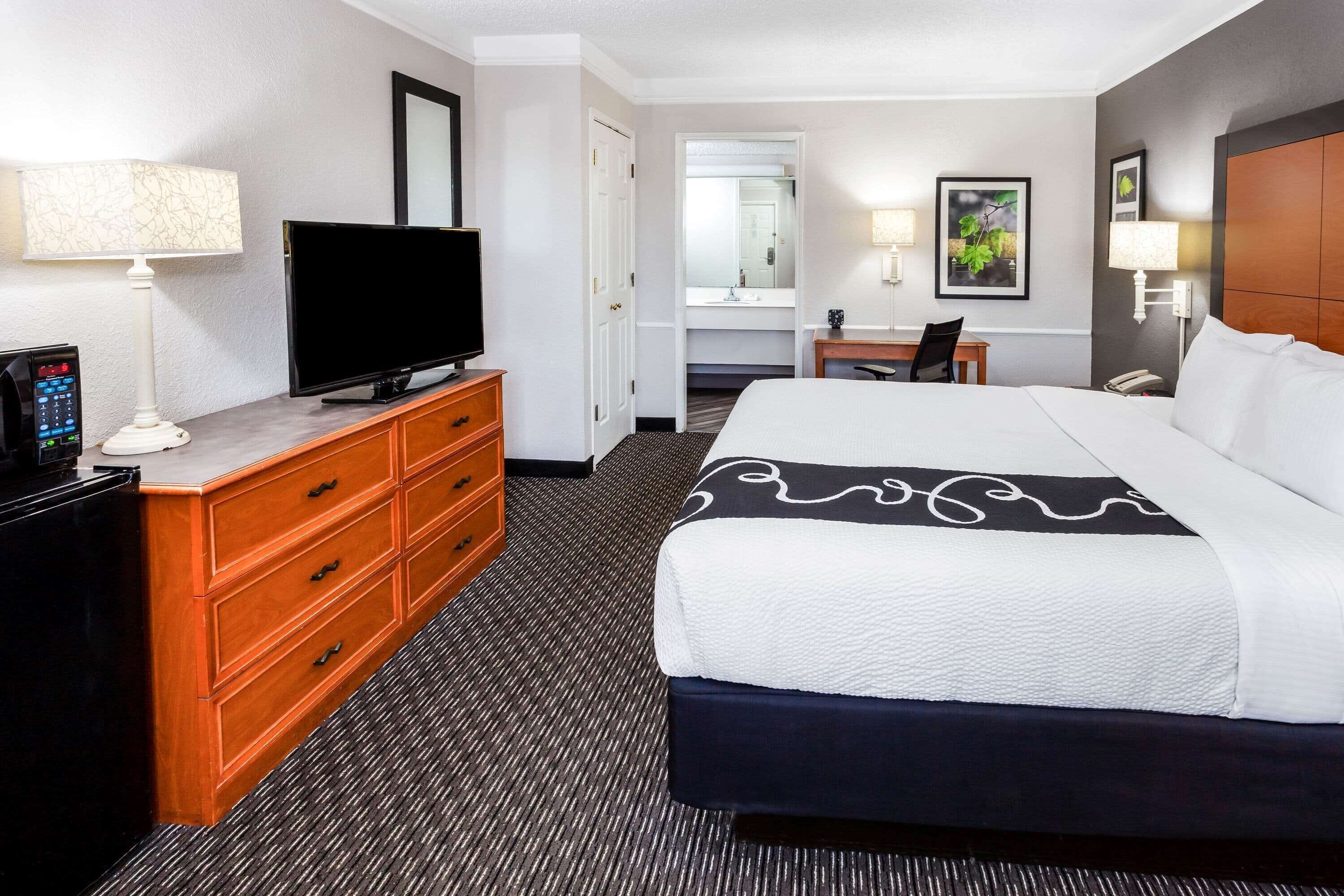 Una habitación de hotel con una cama grande, un televisor sobre un aparador de madera, un escritorio con una silla y un baño visible a través de una puerta abierta.