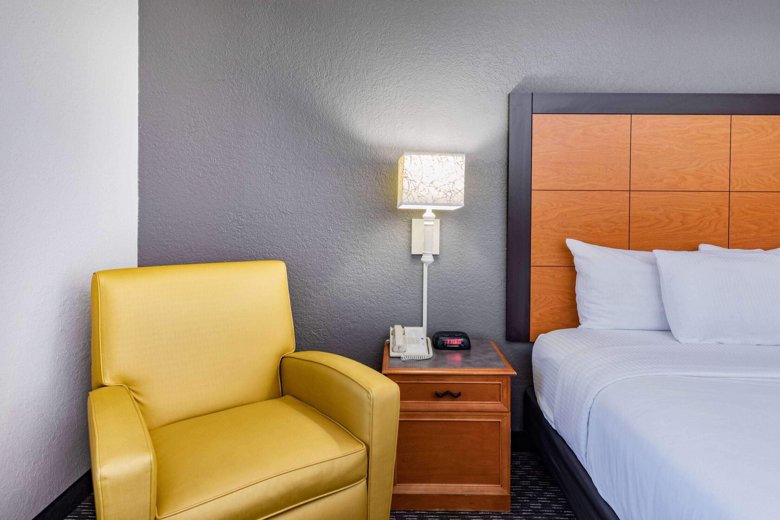A hotel room with a yellow armchair, a bedside table with a lamp, and a bed with white linens.