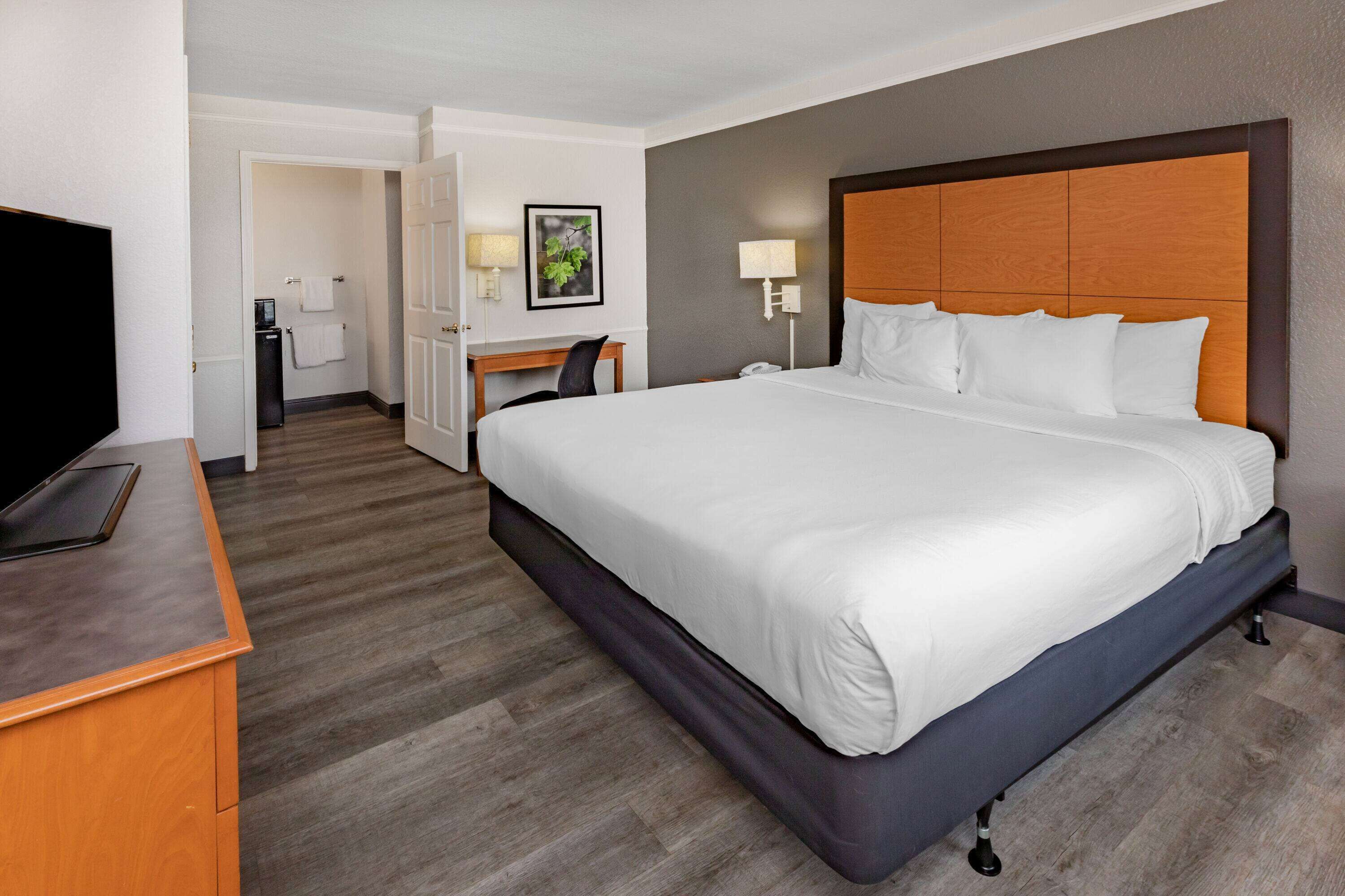 Une chambre d’hôtel avec un grand lit, un bureau, une chaise, une télévision et une salle de bain visible à travers une porte ouverte.