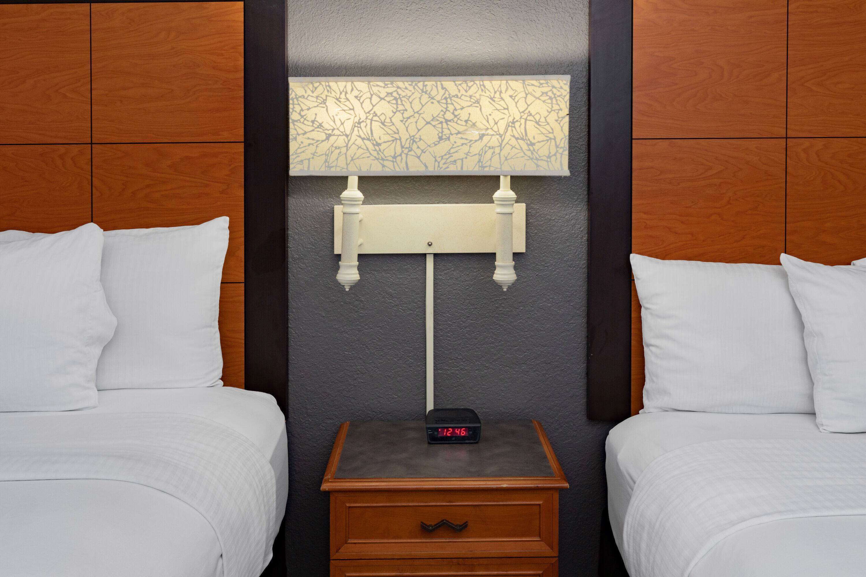 A hotel room with two beds, a nightstand with a clock, and a wall-mounted lamp.