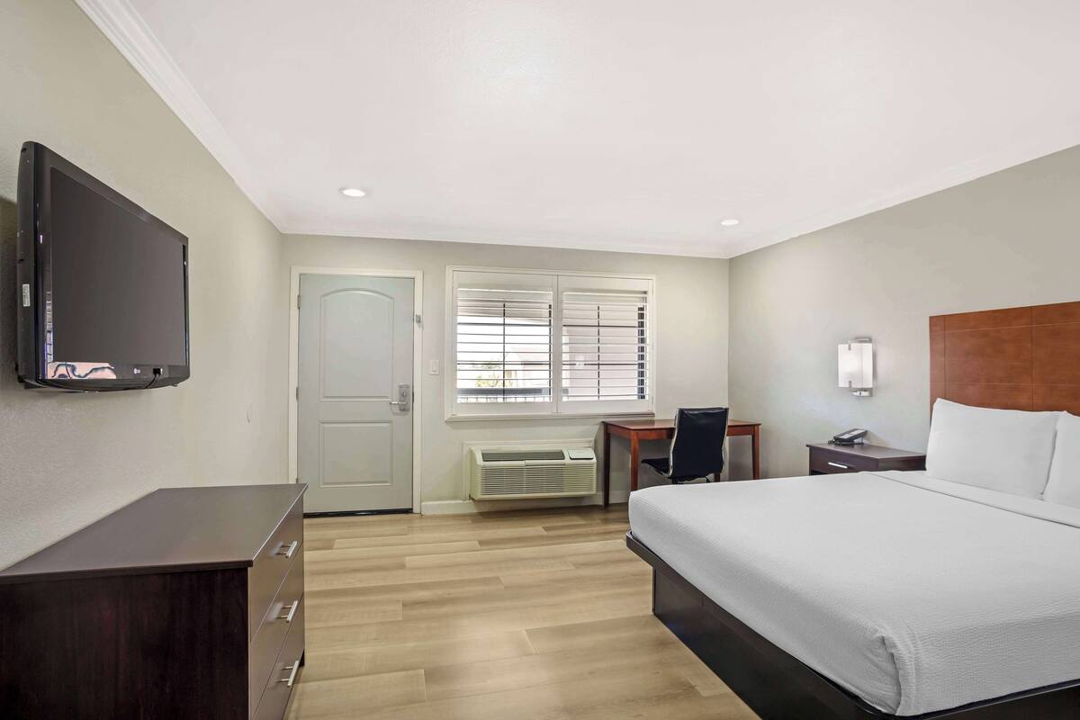 A hotel room with a bed, a desk, a chair, a TV, a dresser, and a window with blinds.