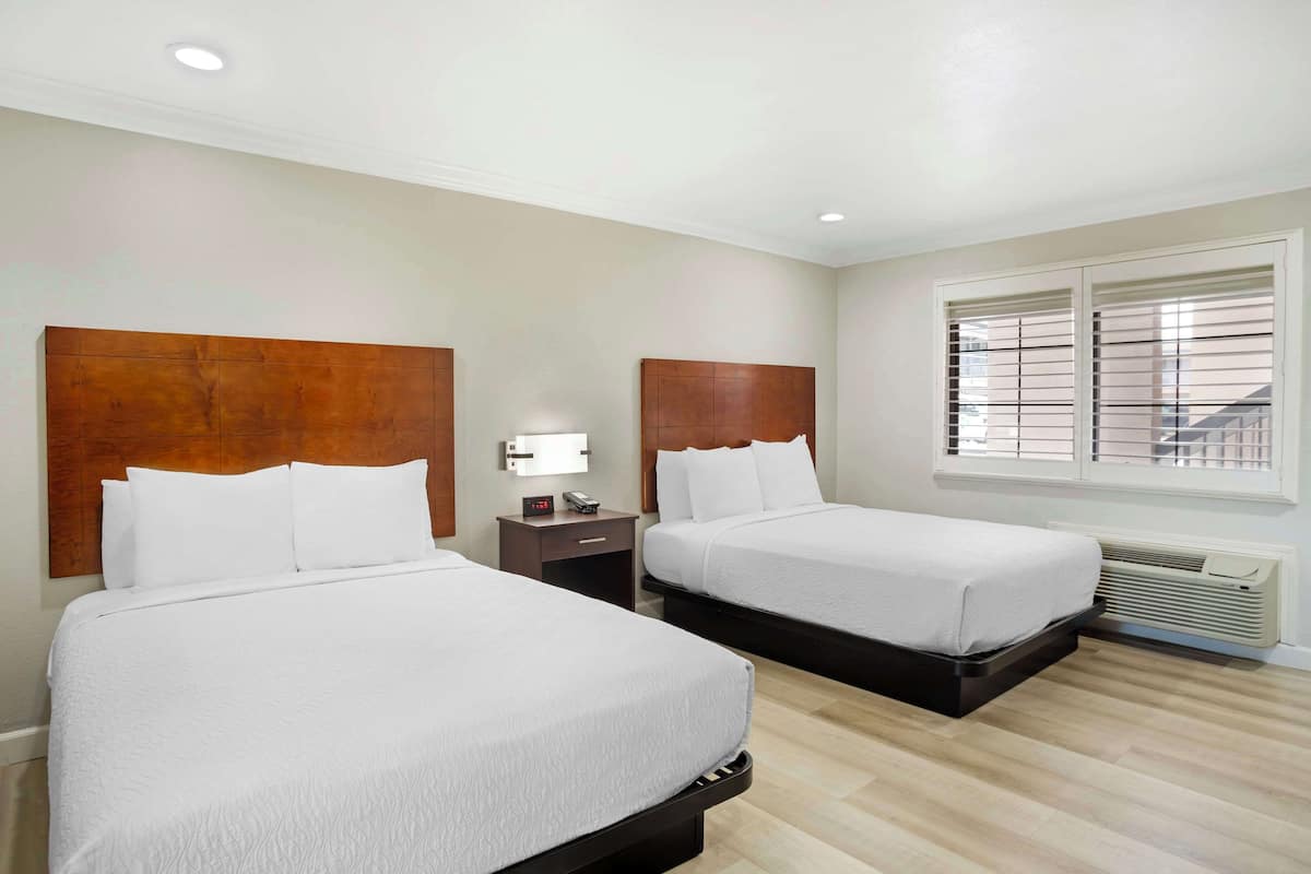A hotel room with two beds, a nightstand with a lamp, a window with blinds, and a wall-mounted heater.