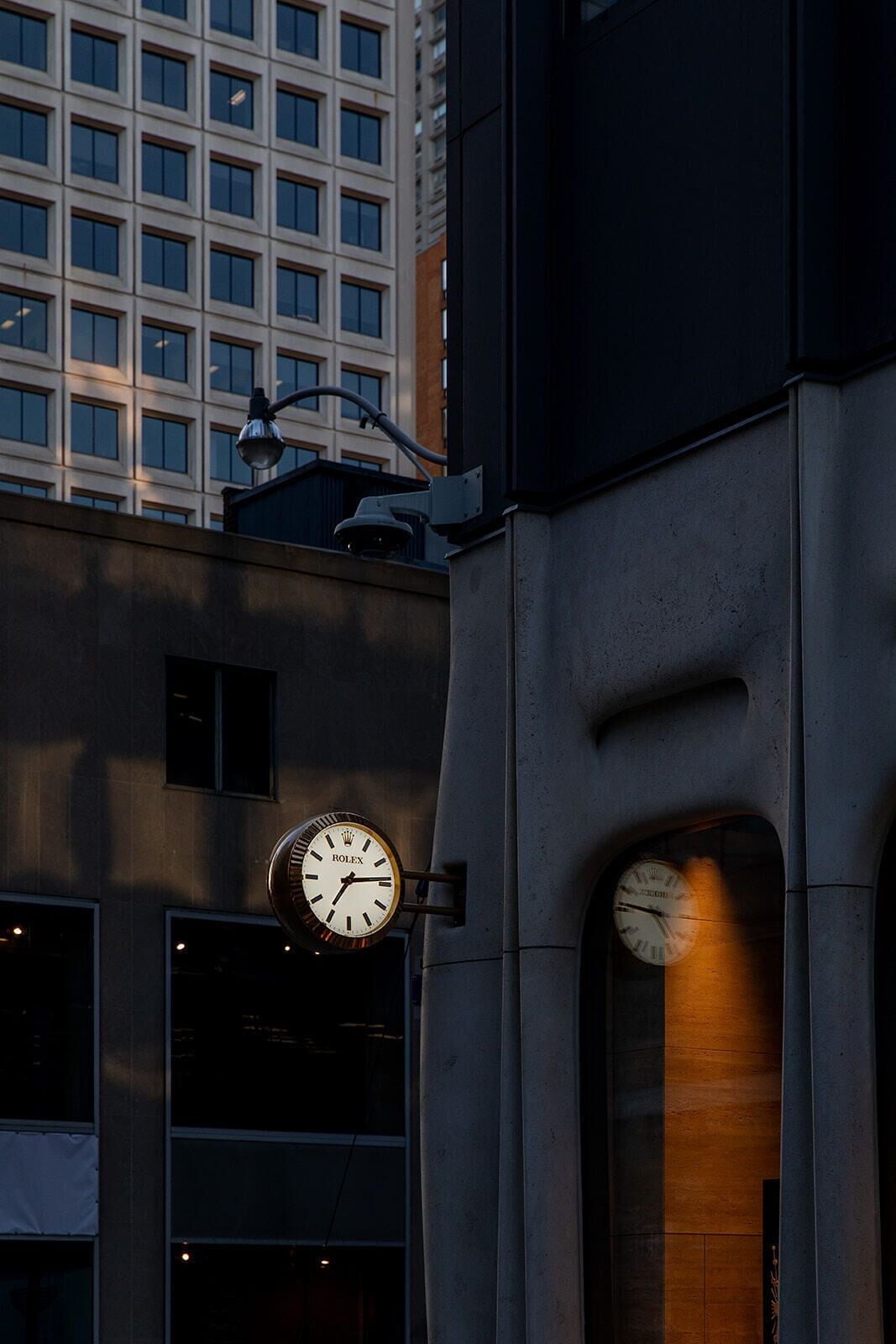 Een gebouw met een Rolex klok aan de buitenmuur, een groot raam en een uitzicht op een hoog kantoorgebouw op de achtergrond.
