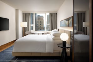 A hotel room with a large bed, a desk, a chair, a TV, and a view of a building through the window.