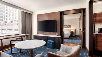 Habitación de hotel moderna con un televisor de pantalla plana grande montado en la pared, una mesa de comedor redonda, un sofá y un pequeño otoman azul.