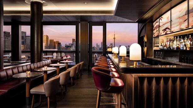 Un bar en la azotea con vistas al horizonte urbano, asientos de cuero y una barra repleta de botellas.