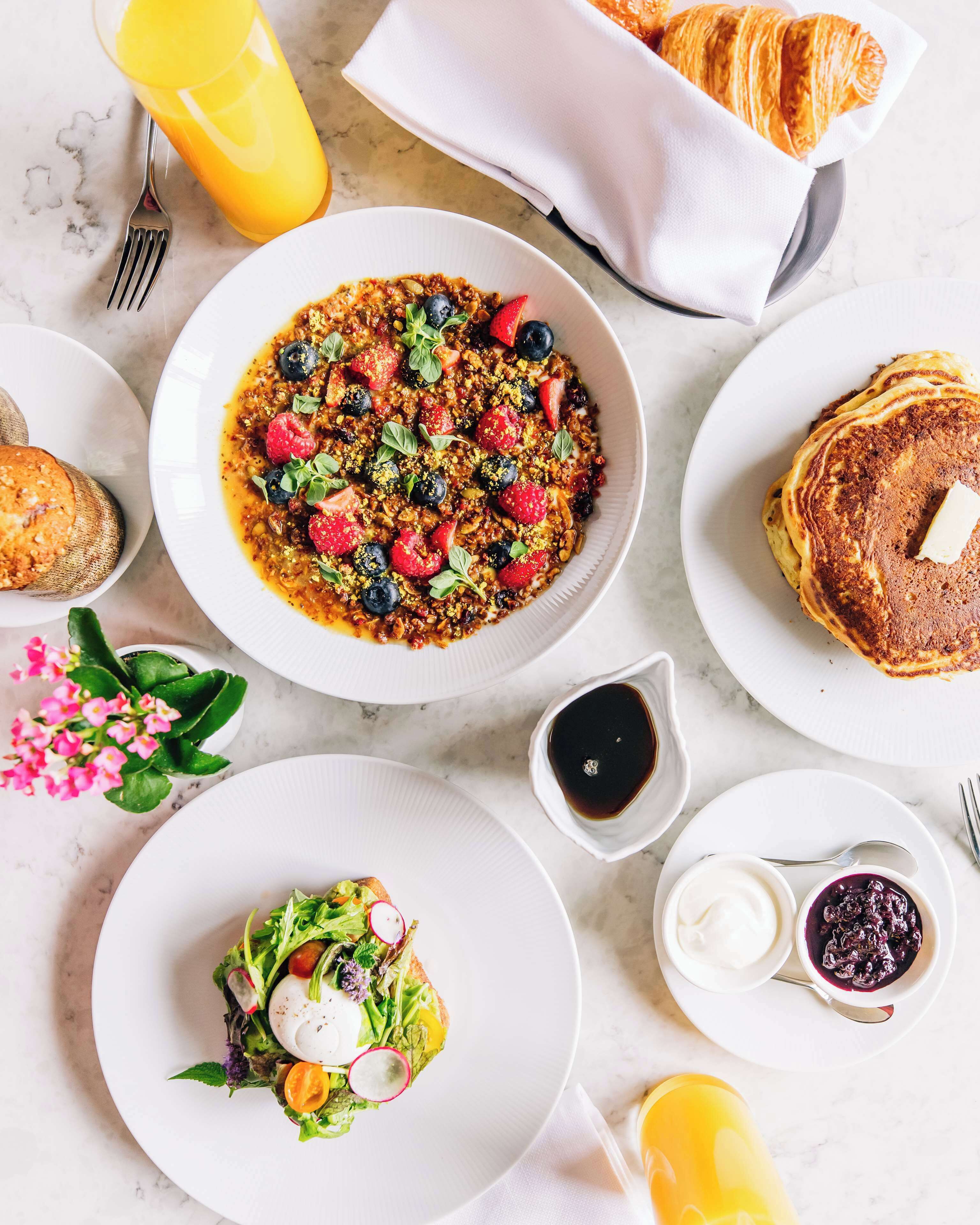 A breakfast setting with a bowl of granola, pancakes, croissants, and a glass of orange juice.