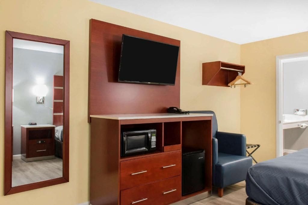 A hotel room with a flat-screen TV mounted on the wall, a wooden cabinet with drawers, a microwave, a small refrigerator, a blue chair, and a bed with a blue cover.