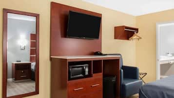 A hotel room with a flat-screen TV mounted on the wall, a wooden cabinet with drawers, a microwave, a small refrigerator, a blue chair, and a bed with a blue cover.