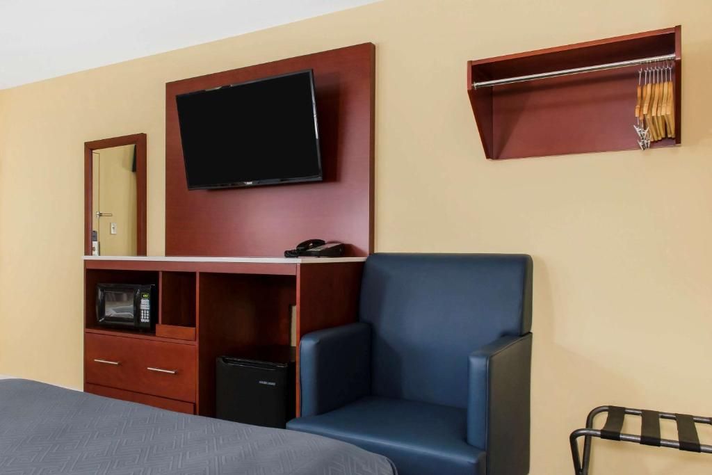 A hotel room with a bed, a TV mounted on the wall, a desk with a microwave, a chair, and a coat rack.