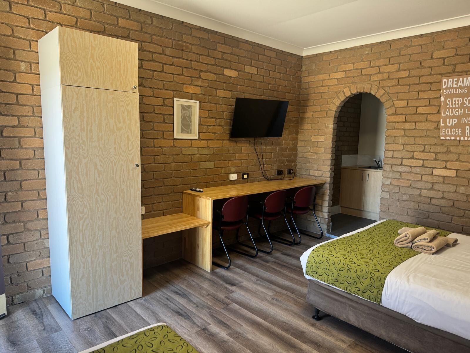 A room with a brick wall, a wooden desk with chairs, a bed with a green patterned bedspread, and a television mounted on the wall.