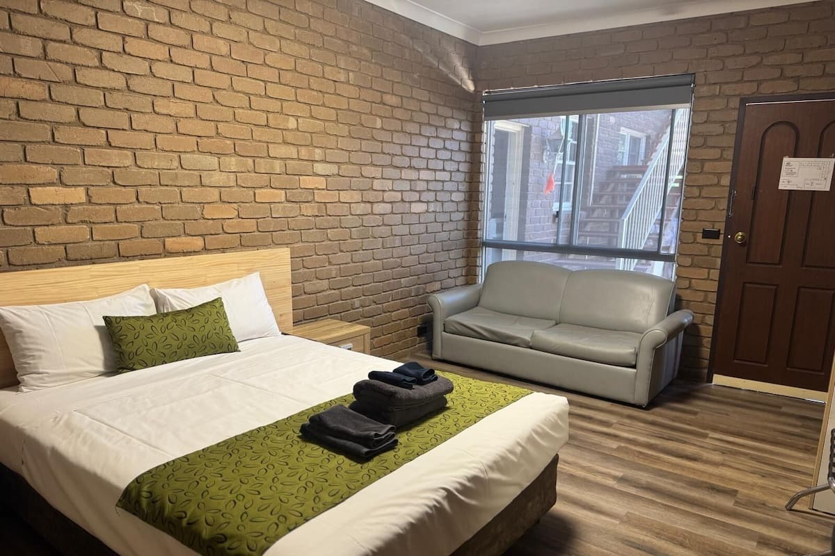 A hotel room with a brick wall, a bed with a green patterned bedspread, a grey sofa, and a wooden floor.