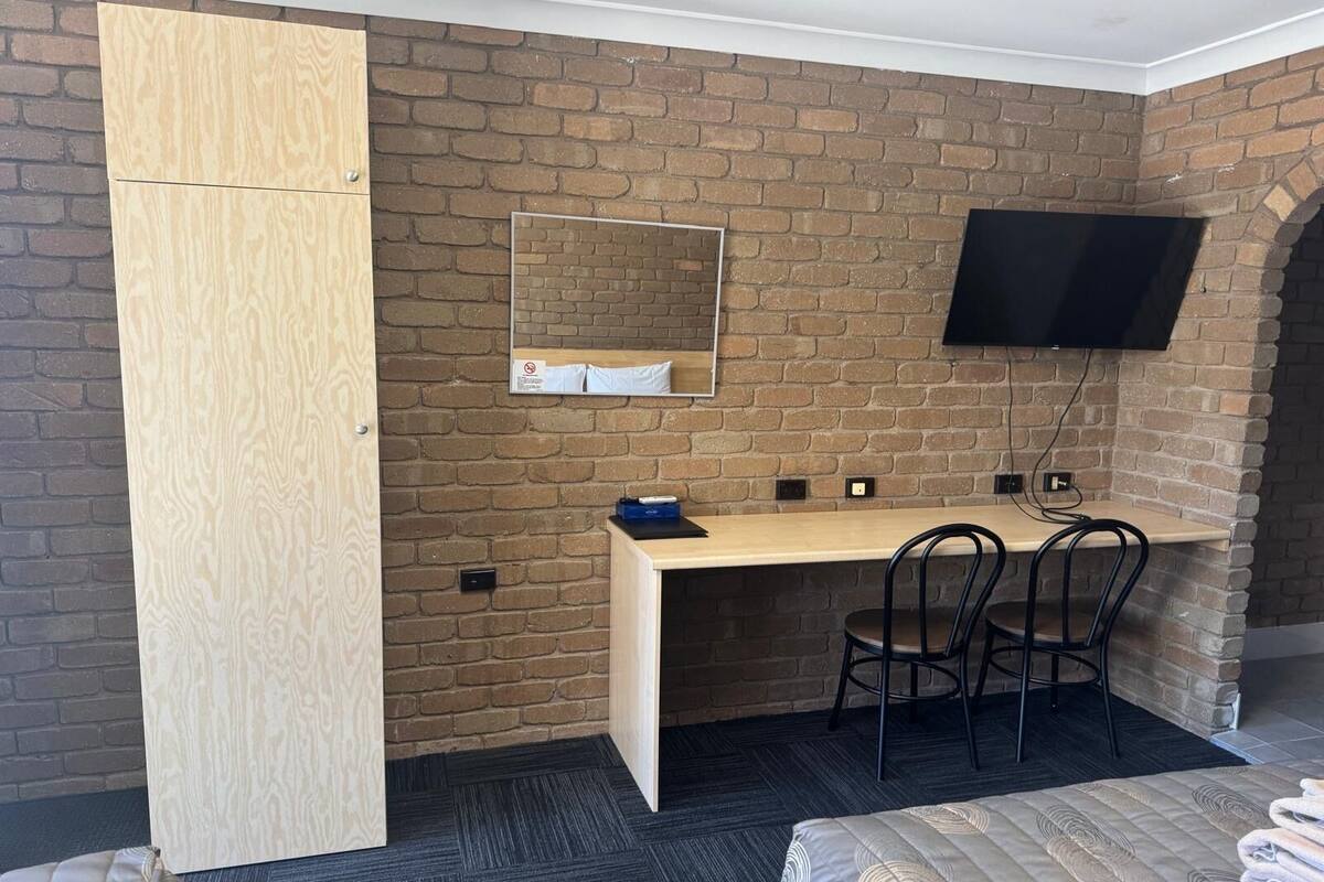 A compact room with a brick wall, a wooden wardrobe, a desk with two chairs, a flat-screen TV mounted on the wall, and a bed with a patterned duvet.