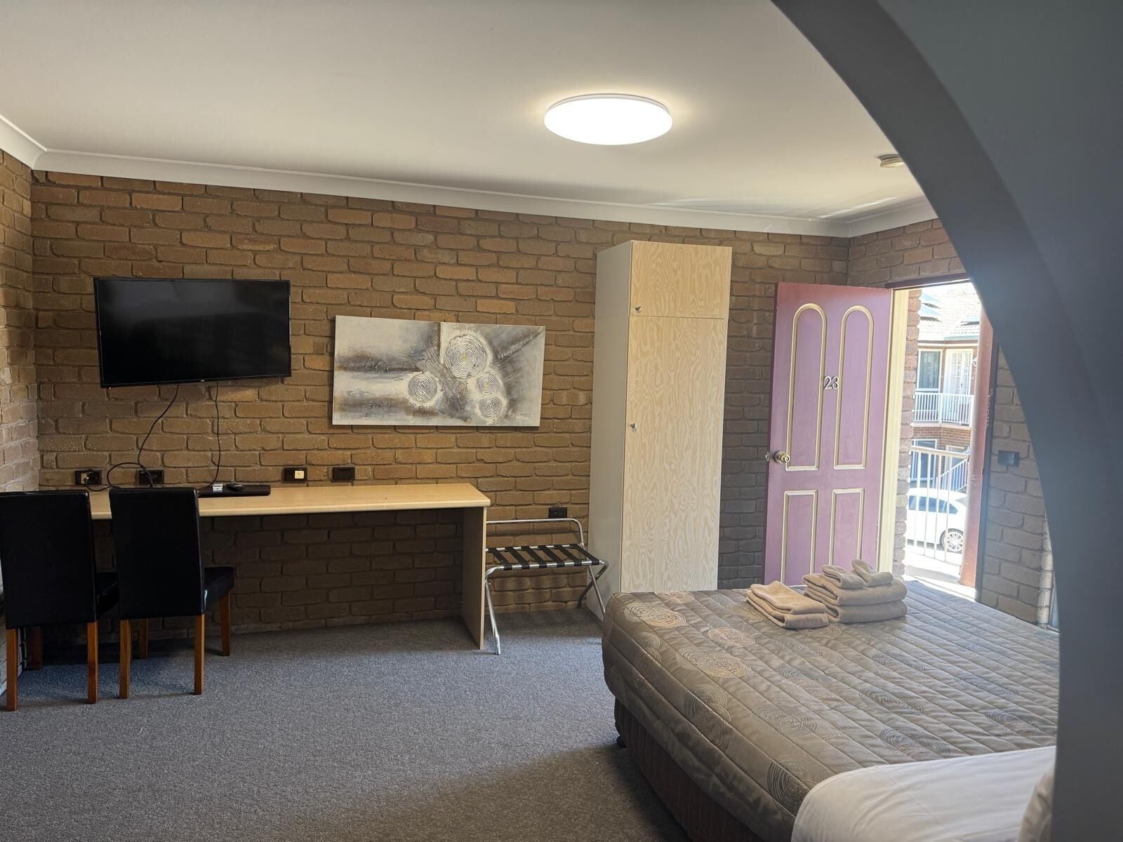 Een kamer met een bakstenen muur, een flatscreen televisie boven een houten bureau, een bed met een gewatteerde spreideken, en een deur met een nummer.