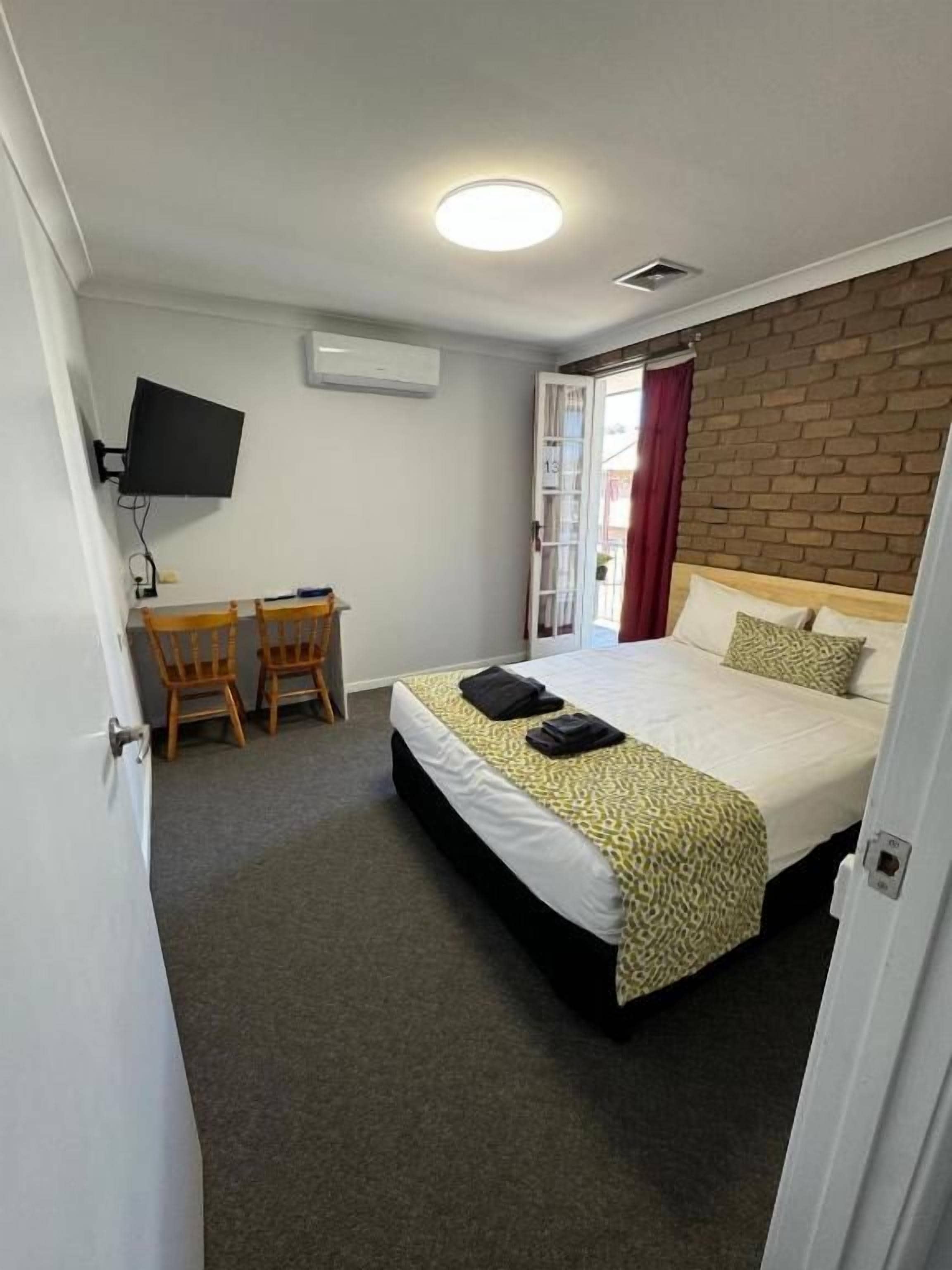 A hotel room with a bed, a desk with two chairs, a television, and a brick wall.