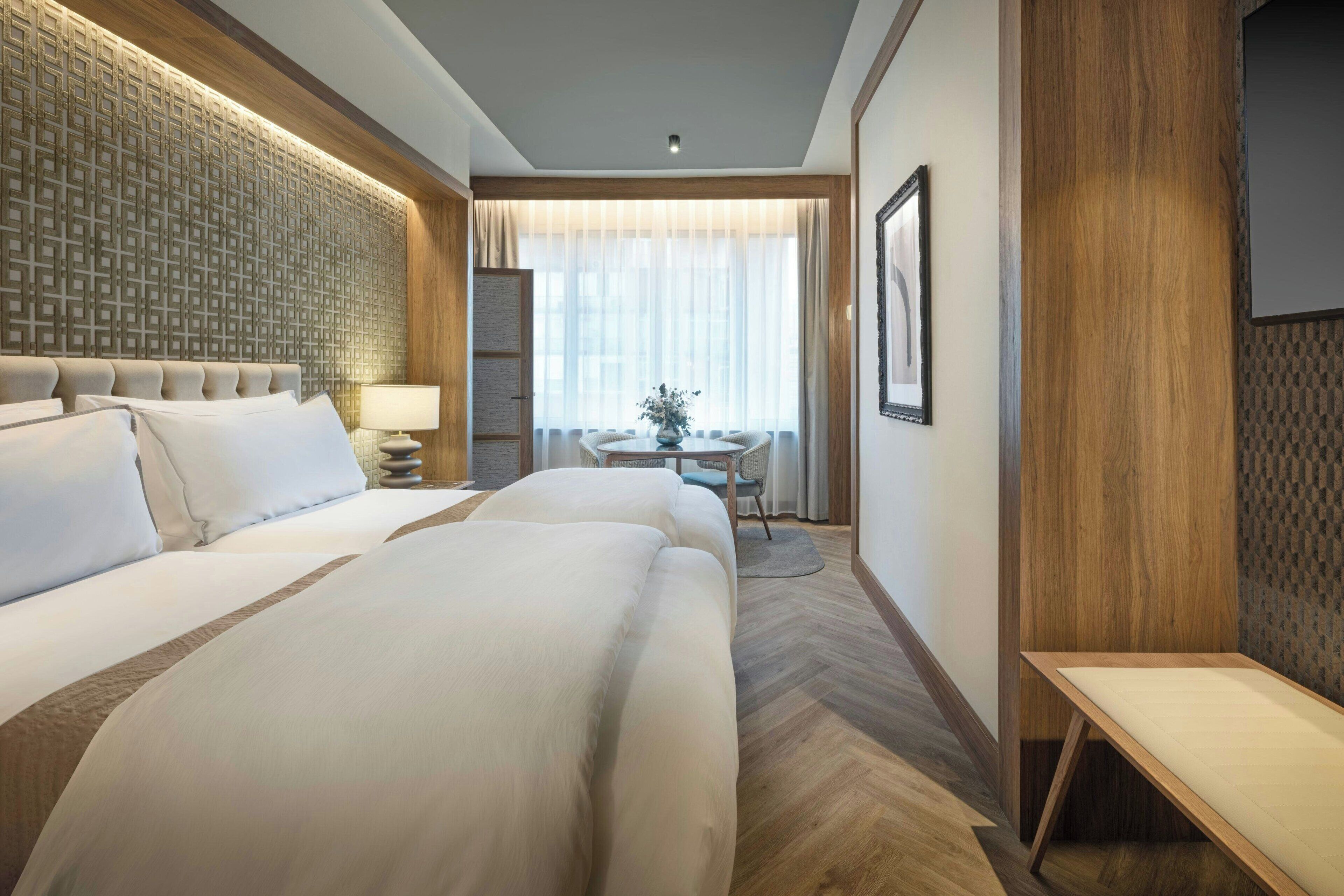 A hotel room with two large beds, a wooden headboard, a small table with a chair, and a large window with sheer curtains.