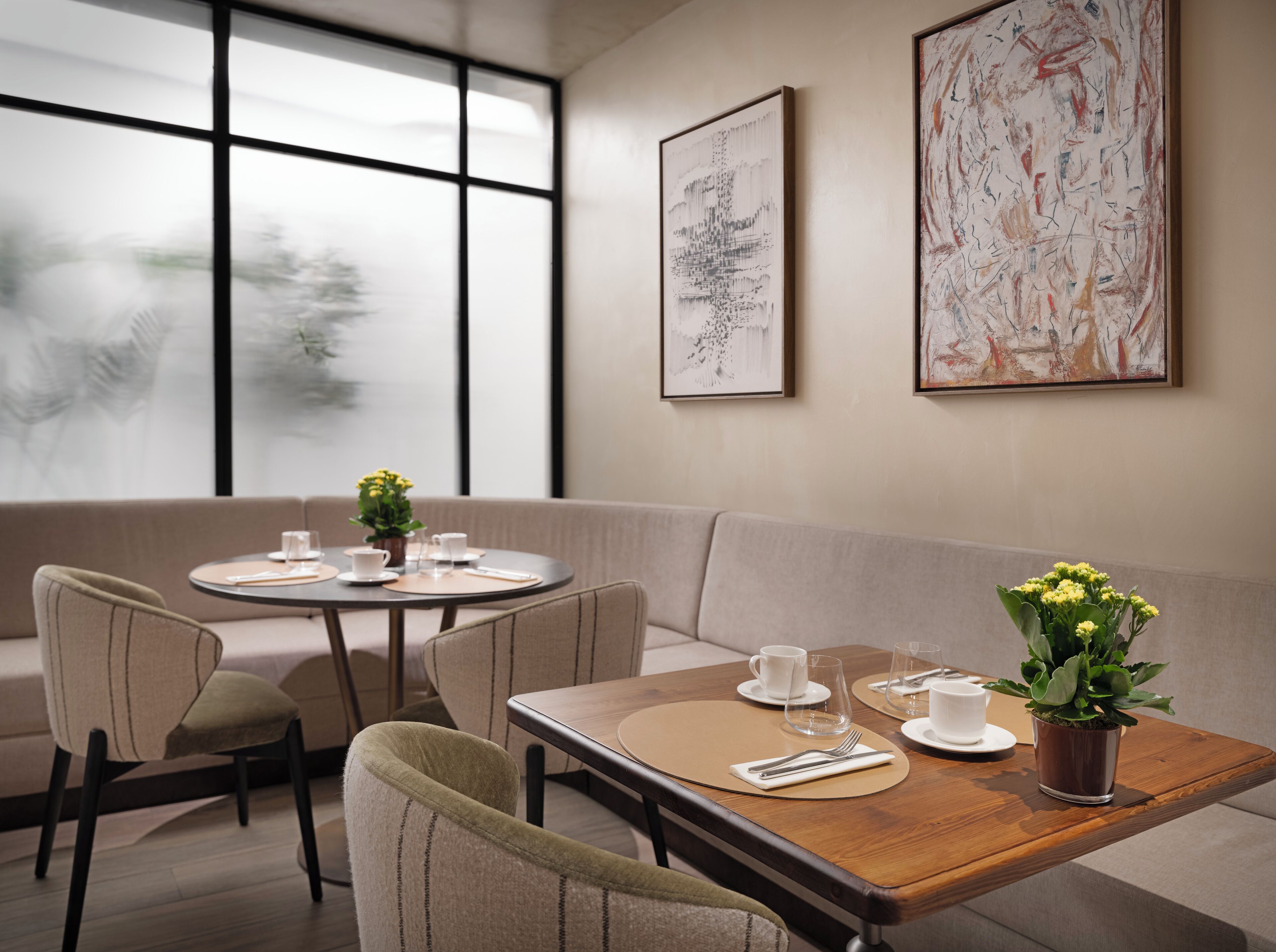 A modern dining area with a round table, chairs, and a sofa. There are framed artworks on the wall and a window with a view.