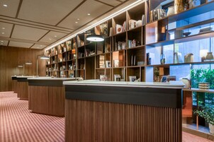 Eine Hotellobby mit einem hölzernen Empfangstresen, einem Bücherregal mit Büchern und Dekorationsgegenständen sowie Blick auf Grünflächen.