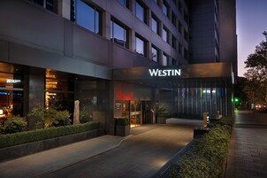 Ein moderner Hotelzugang mit einem gut sichtbaren 'WESTIN'-Schild, einem gut beleuchteten Weg und einer angelegten Grünfläche mit Büschen und einem Steinobjekt.