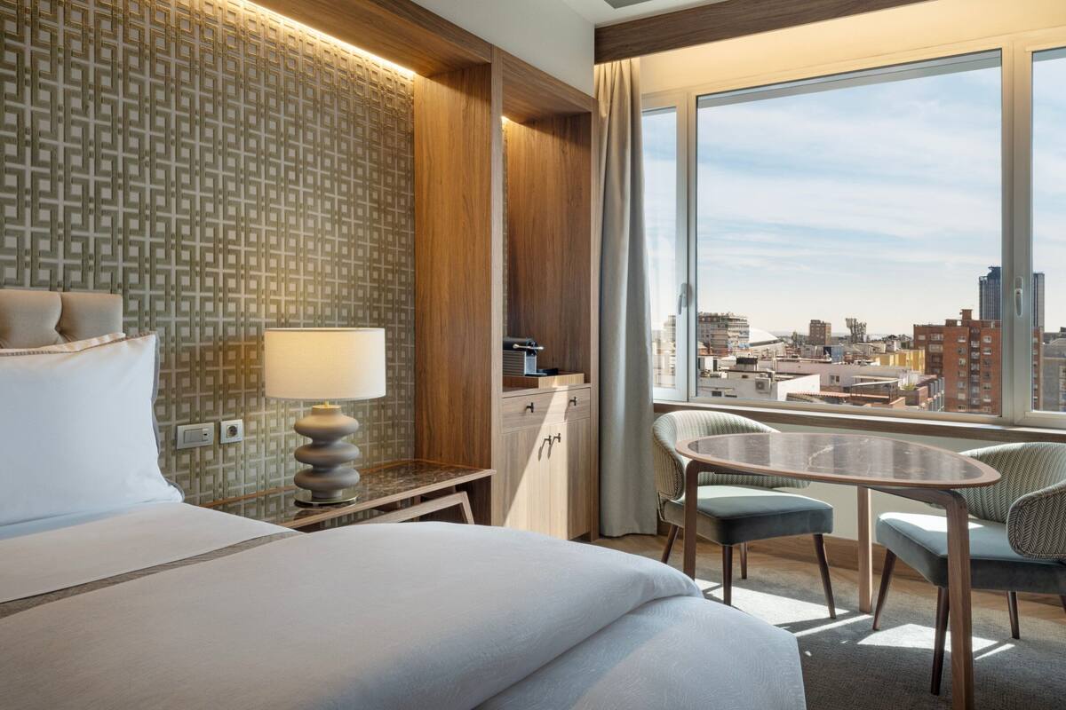 Une chambre d’hôtel avec un grand lit, un bureau avec une lampe, une chaise, une petite table et une vue sur la ville grâce à de grandes fenêtres.
