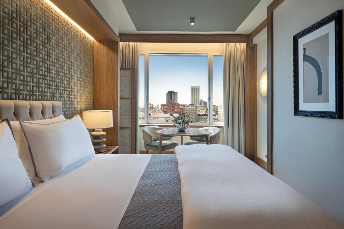 Une chambre d’hôtel avec un grand lit, une petite table avec des chaises, une vue sur la ville et un panneau décoratif au mur.