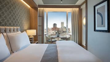 Habitación de hotel con una cama grande, una mesita con sillas, vista a la ciudad y un panel decorativo en la pared.