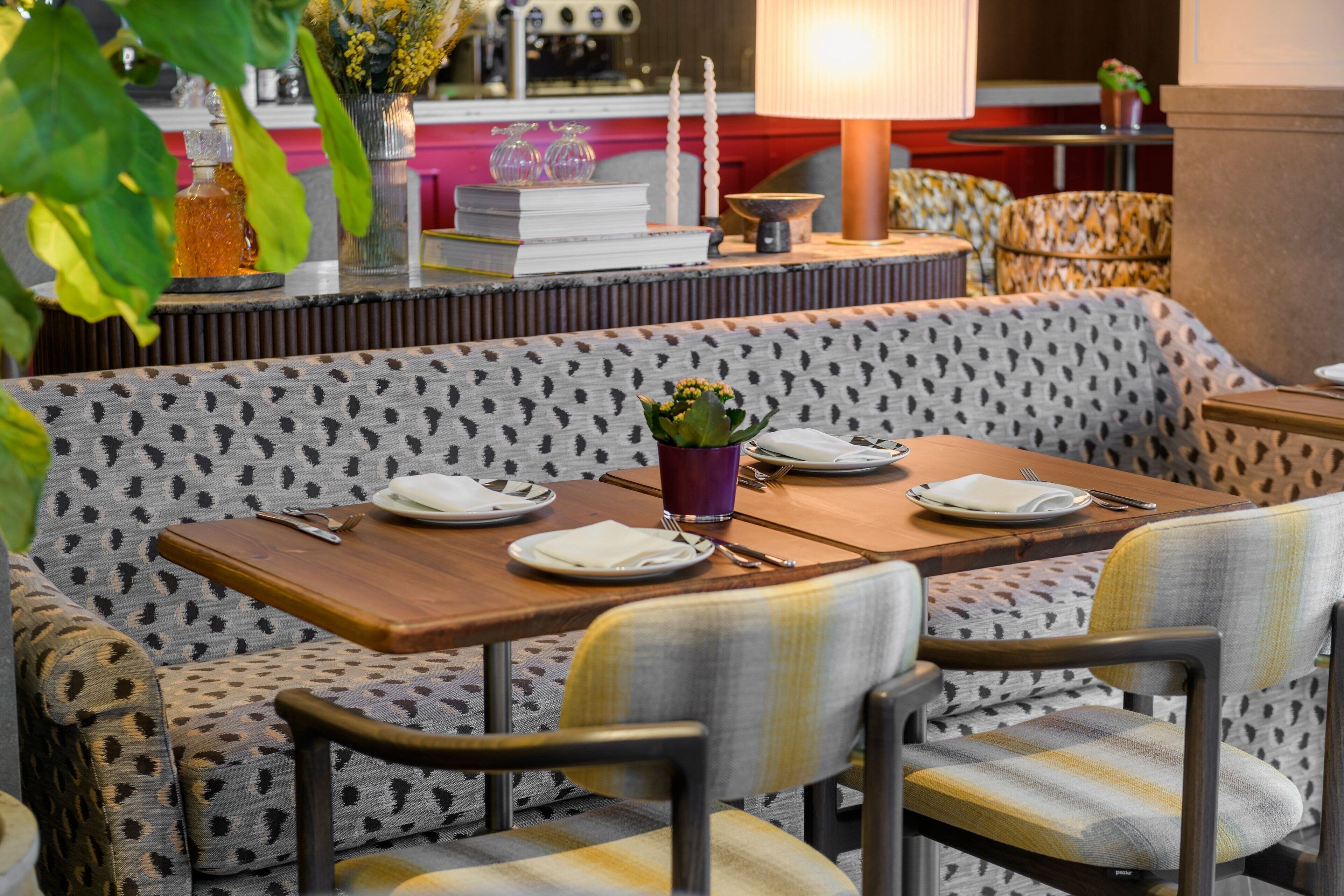 A dining area with a patterned sofa, a wooden table set for two, and a vase with flowers.