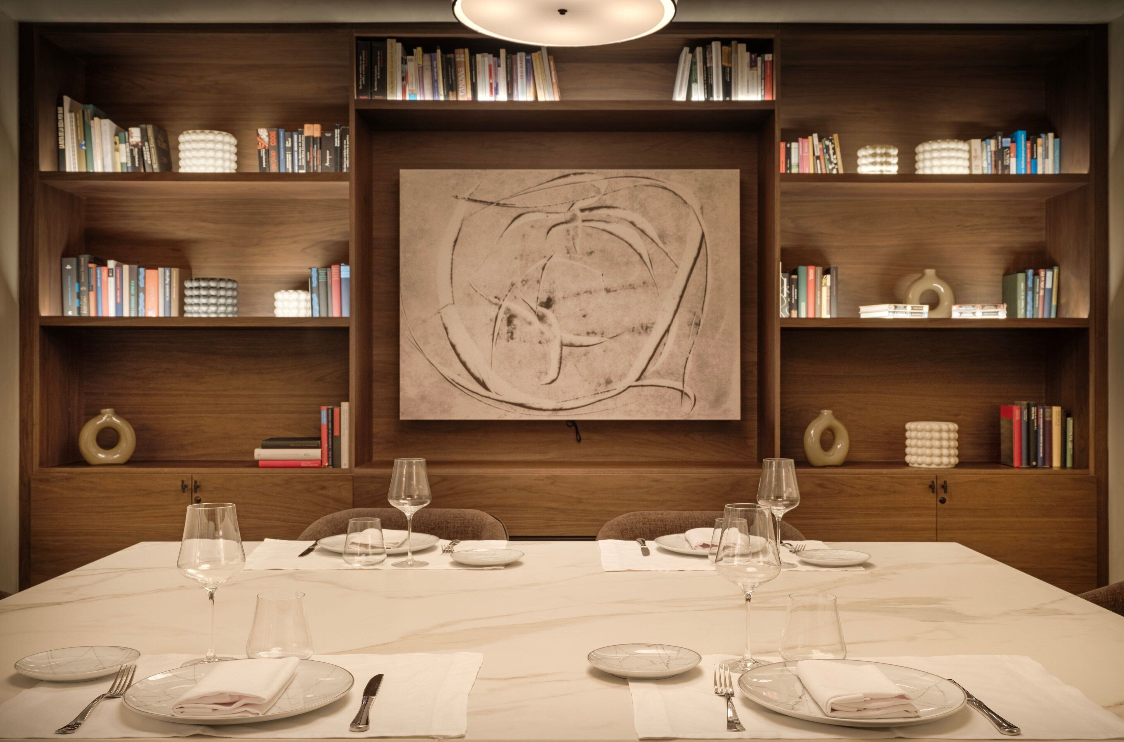 Mesa de comedor dispuesta para cuatro, con copas de vino y platos, flanqueada por estanterías llenas de libros y objetos decorativos.