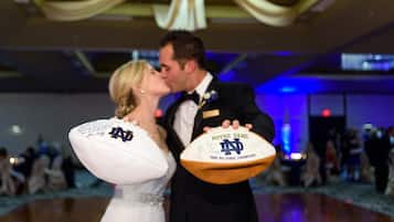 A couple holding footballs with Notre Dame logos, in a room with a dance floor and people in the background.
