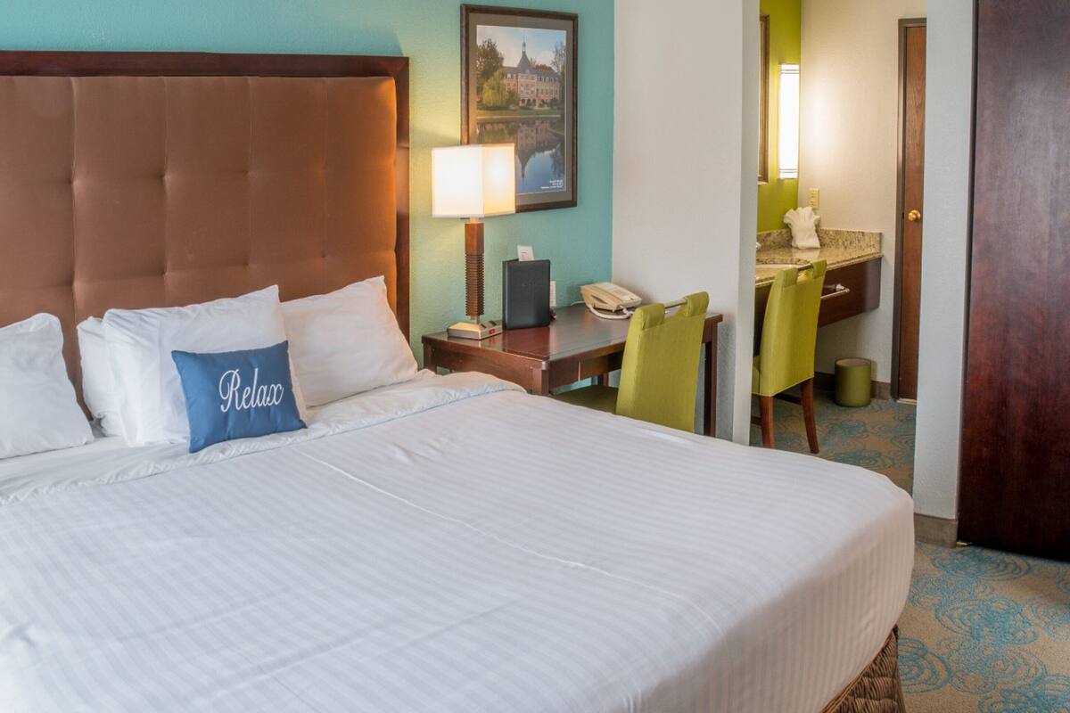 A hotel room with a large bed, a nightstand with a lamp, a desk with a telephone, and a chair.