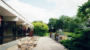An outdoor patio with tables and chairs, surrounded by greenery and a building with a glass facade.
