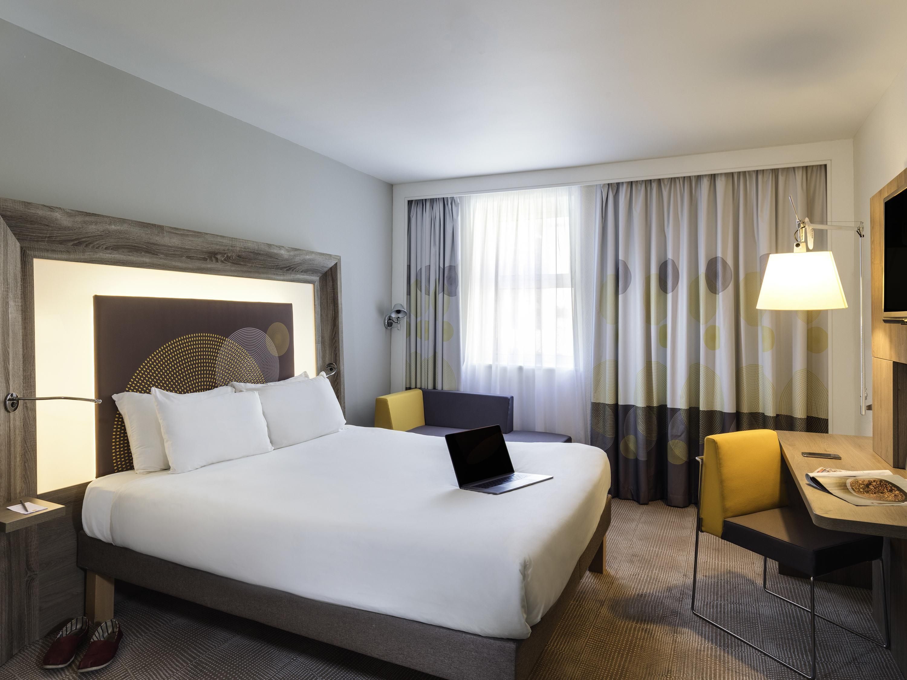 A hotel room with a large bed, a desk, a chair, a laptop, and a TV.