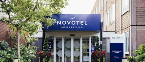 The Novotel hotel entrance with a glass door, a signboard, and potted plants.