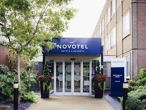 The Novotel hotel entrance with a glass door, a signboard, and potted plants.