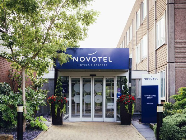 The Novotel hotel entrance with a glass door, a signboard, and potted plants.