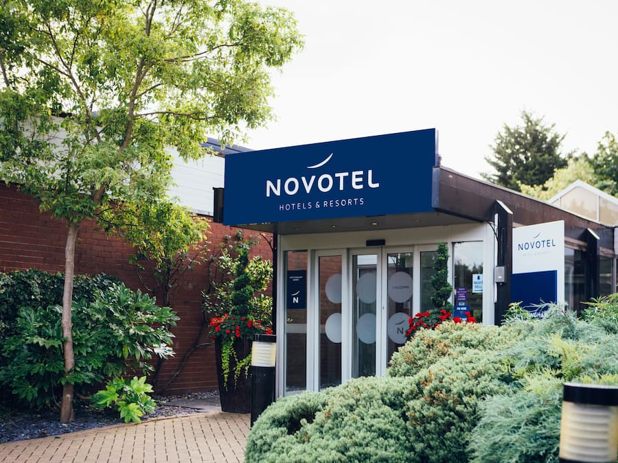 A Novotel hotel entrance with a glass door, surrounded by greenery and a brick wall.