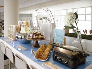 A buffet counter with a variety of food items, a waffle plate, a waffle iron, and a decorative vase with flowers.