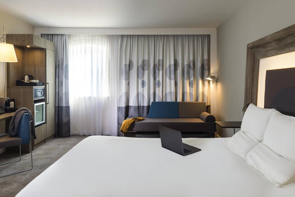 A hotel room with a bed, a desk with a laptop, a sofa, a coffee machine, and a TV.