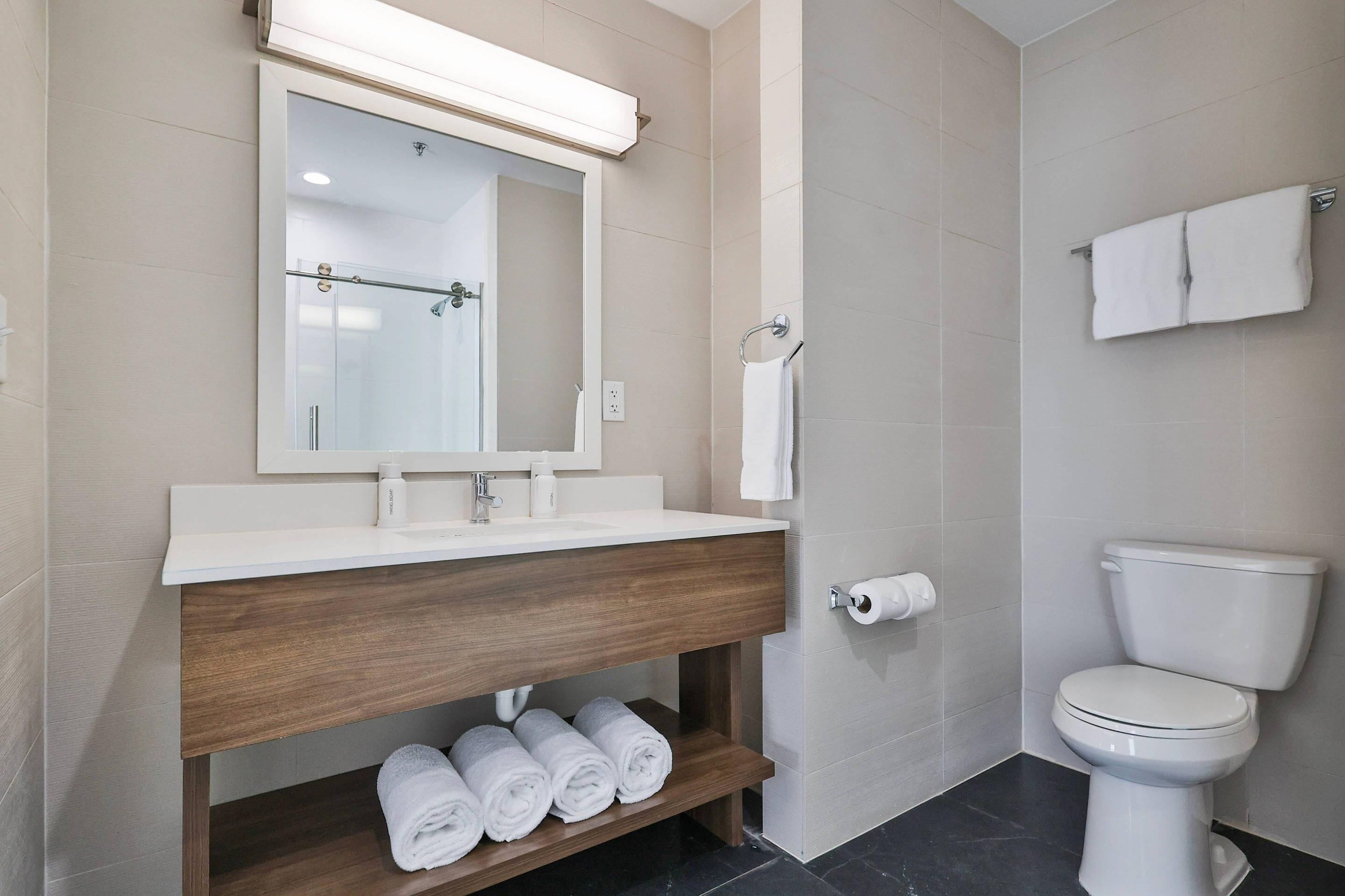 Un baño moderno con lavamanos, espejo y retrete blancos. Hay toallas en un estante y un perchero en la pared.