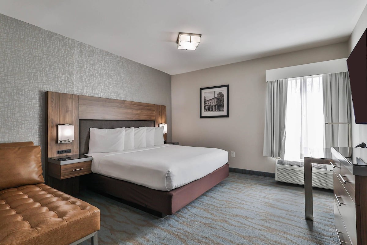 A hotel room with a large bed, a wooden headboard, a bedside table with a lamp, a leather chair, and a large window with curtains.