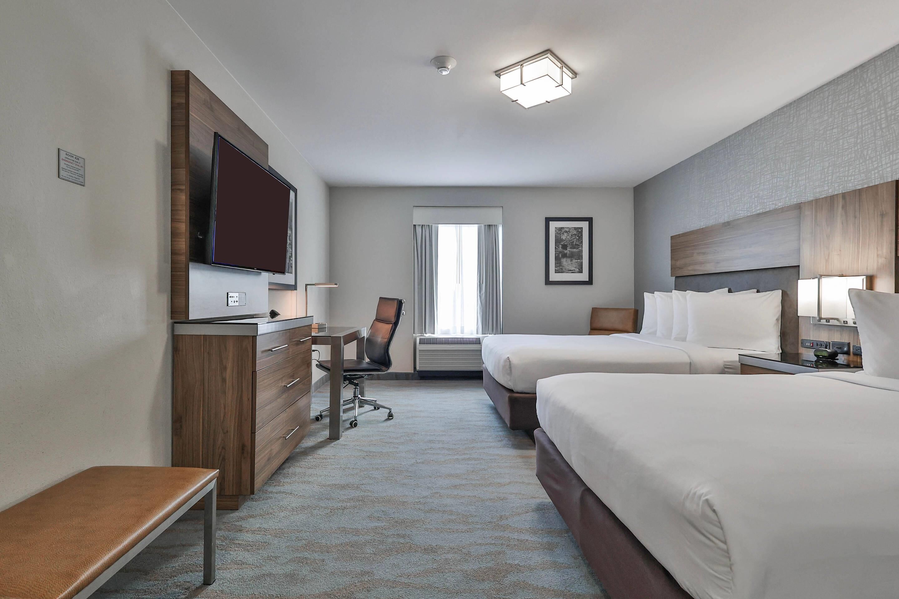 A hotel room with two beds, a desk with a chair, a flat-screen TV, a bench, and a window with curtains.