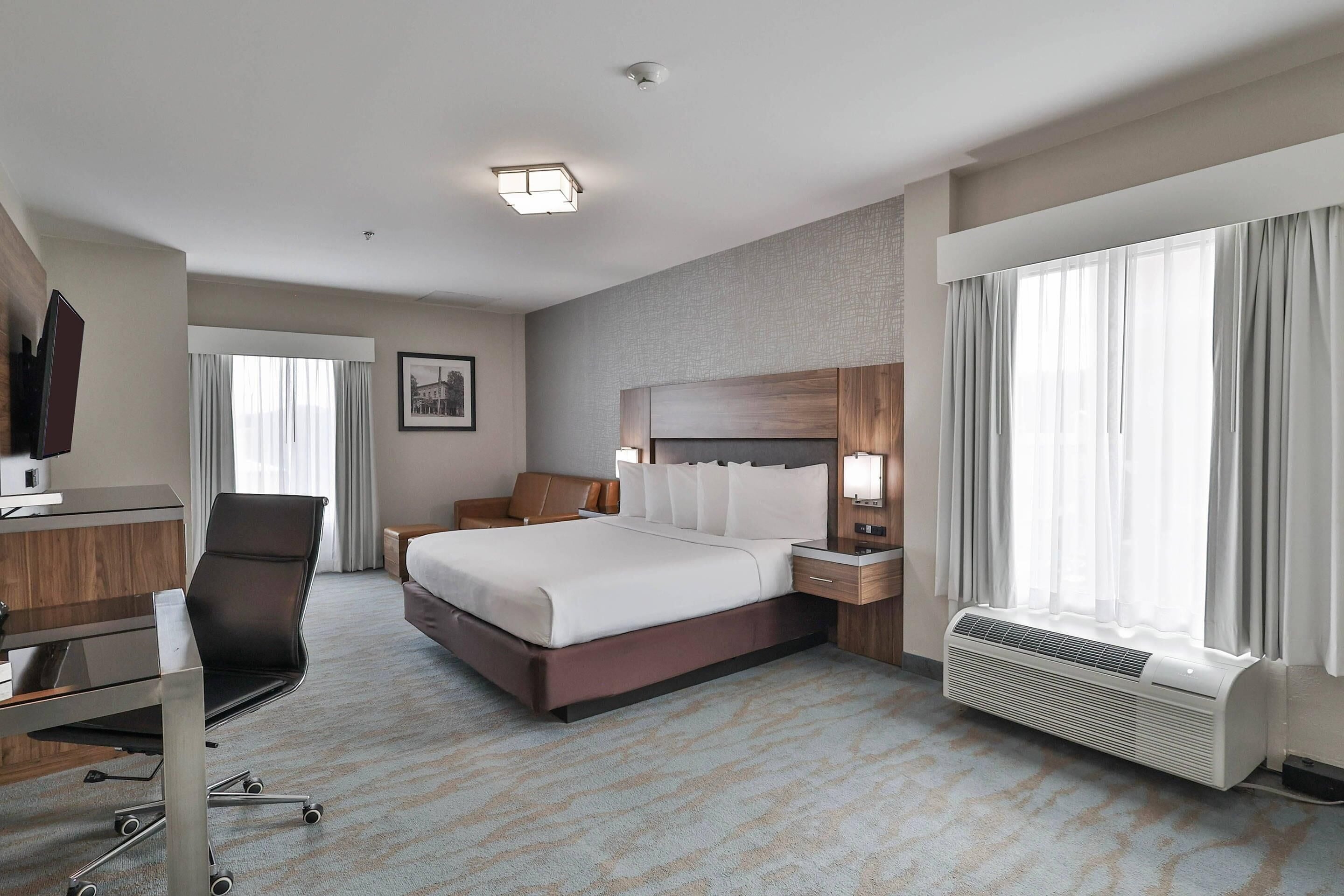 A hotel room with a large bed, a desk, a chair, a TV, a bedside table with a lamp, a window with curtains, and a wall-mounted air conditioner.