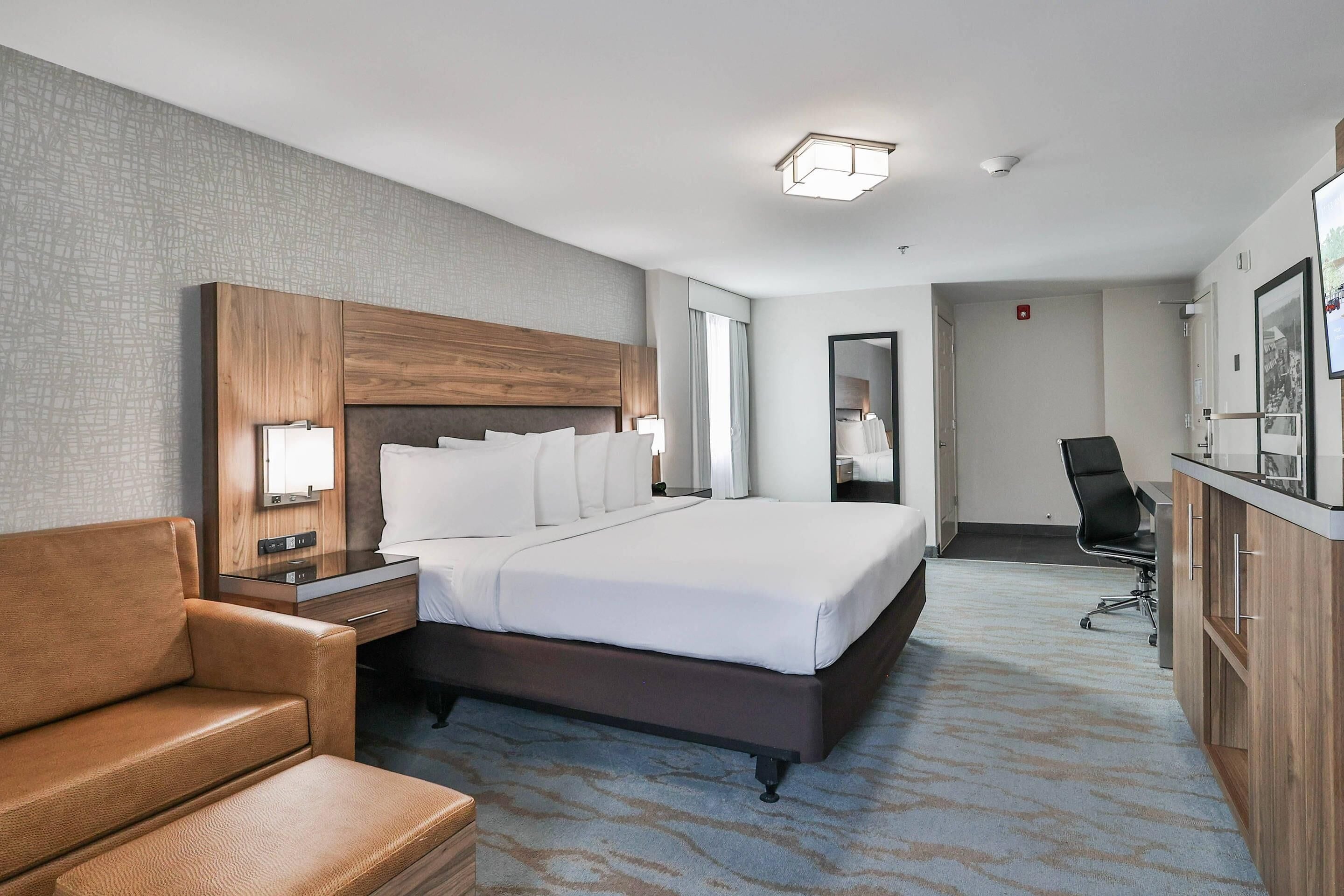 A hotel room with a large bed, a wooden headboard, a leather armchair, a desk with a chair, and a minibar.
