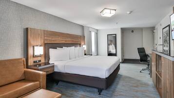 A hotel room with a large bed, a wooden headboard, a leather armchair, a desk with a chair, and a minibar.