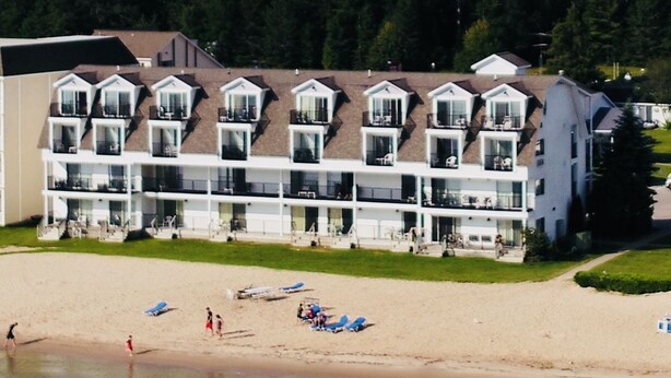 A multi-story hotel with balconies, a beach in the foreground, and a forested hill in the background. - Quality Inn & Suites Mackinaw City Beachfront (Mackinaw City)
